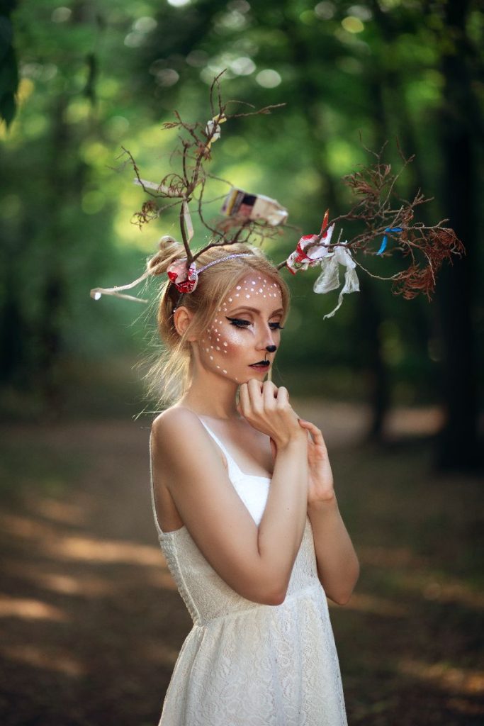 Szőke hajú nő őzikének sminkelve áll az erdőben, fehér ruhában. A fején száraz ágak imitálják az agancsokat, amikre hulladék darabok vannak felakadva